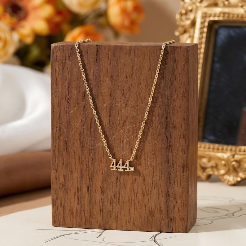 May include: A gold-colored necklace with the number "444" pendant. The necklace is displayed on a wooden block. The background includes a gold-framed mirror and orange flowers.