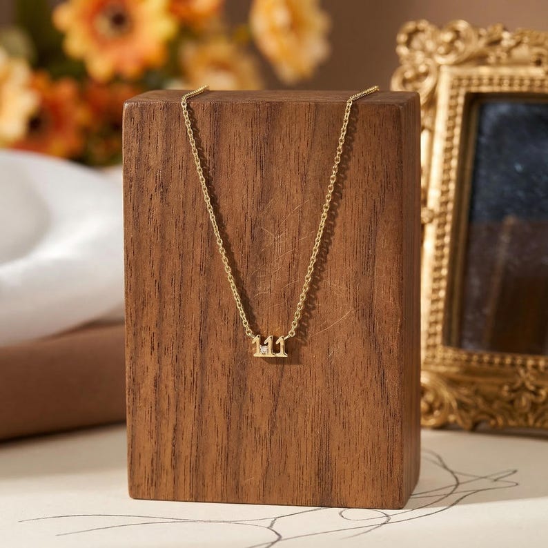 May include: A delicate gold-colored necklace with the number 111 pendant. The necklace is displayed on a wooden block, with a blurred background featuring flowers and a decorative gold frame. The pendant has a small, sparkling accent.