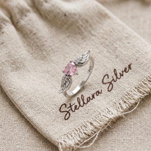 May include: A silver ring featuring a pink heart-shaped gemstone and wing detailing. The ring is displayed on a beige fabric, with the words "Stellara Silver" in brown script.