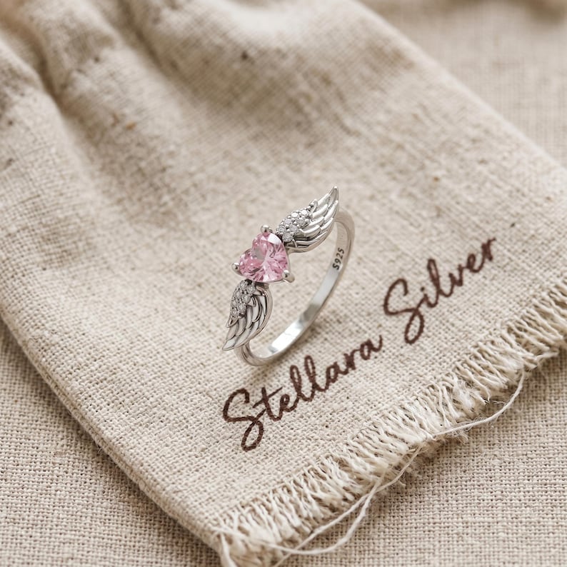 May include: A silver ring featuring a pink heart-shaped gemstone and wing detailing. The ring is displayed on a beige fabric, with the words "Stellara Silver" in brown script.