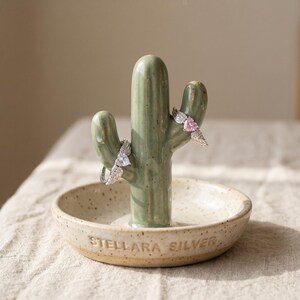 May include: A ceramic cactus ring holder in a speckled beige dish with the text "STELLARA SILVER." Two rings with wing designs are displayed on the cactus. The cactus is a muted green colour.