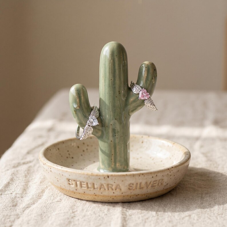 May include: A ceramic cactus ring holder in a speckled beige dish with the text "STELLARA SILVER." Two rings with wing designs are displayed on the cactus. The cactus is a muted green colour.