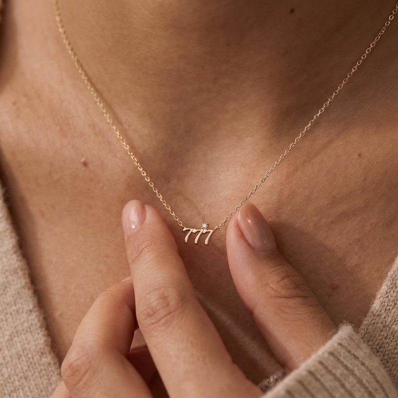 May include: A delicate gold necklace featuring the number "777" with a small gemstone accent. The pendant hangs from a fine chain, creating a minimalist and elegant design. The necklace is shown on a person's neck.