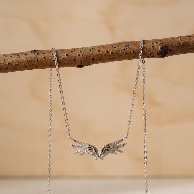 May include: A silver-colored necklace featuring a pair of detailed angel wings. The wings are the focal point, with intricate feather detailing. The necklace hangs from a delicate chain, displayed against a neutral background.