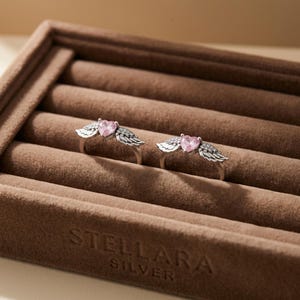 May include: Two silver rings, each featuring a pink heart-shaped gemstone and wing detailing. The rings are presented in a brown velvet jewellery box. The box is embossed with the text "STELLARA SILVER".