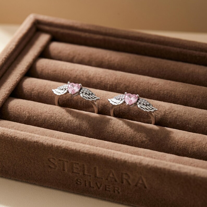 May include: Two silver rings, each featuring a pink heart-shaped gemstone and wing detailing. The rings are presented in a brown velvet jewellery box. The box is embossed with the text "STELLARA SILVER".