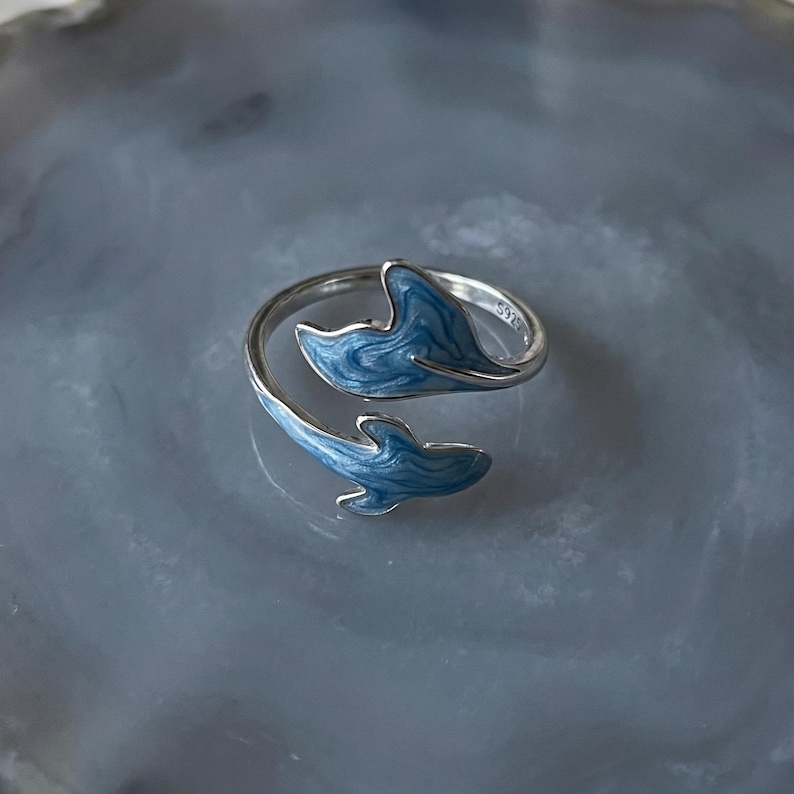 May include: A silver-colored ring with a wrap-around design featuring two blue whale tail shapes. The whale tails have a textured, wave-like pattern and are set against a gray background.