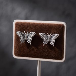 May include: A pair of silver-toned butterfly-shaped stud earrings featuring intricate engraved wing patterns. The earrings are displayed side-by-side on a rectangular brown velvet-textured card with a metallic silver border and a thin metal stand.