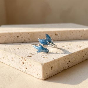 May include: A silver ring with two dolphin tail designs. The tails are painted with a blue and white swirl pattern. The ring is displayed on a textured, light-colored surface.