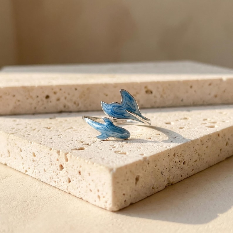 May include: A silver ring with two dolphin tail designs. The tails are painted with a blue and white swirl pattern. The ring is displayed on a textured, light-colored surface.