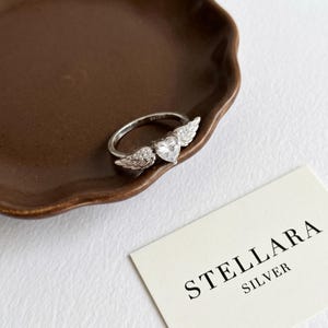 May include: A silver ring featuring a heart-shaped gemstone and wing detailing. The ring sits on a brown plate, with a card reading "STELLARA SILVER". The ring's design includes a central heart-shaped stone and intricate wing embellishments.