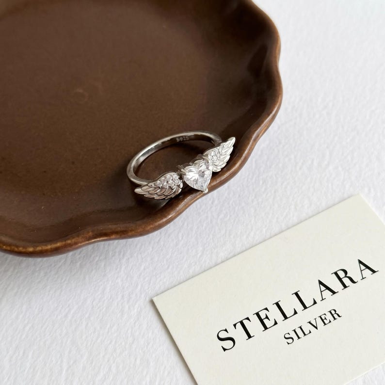 May include: A silver ring featuring a heart-shaped gemstone and wing detailing. The ring sits on a brown plate, with a card reading "STELLARA SILVER". The ring's design includes a central heart-shaped stone and intricate wing embellishments.