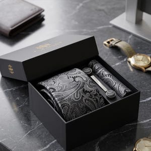Könnte beinhalten: Eine schwarze Geschenkbox mit einer grau-schwarzen Krawatte mit Paisleiderkrawatte mit Paisleidung mit Paisley-Muster, einem passenden Einstecktuch, einer silberfarbenen Krawattenklammer und zwei runden Manschettenknöpfen. Der Deckel trägt ein goldenes Logo und ist geöffnet.