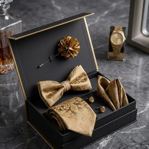 Könnte beinhalten: Eine schwarze Geschenkbox mit einer goldenen Krawatte mit Paisley-Muster, einer passenden Fliege, einem Einstecktuch, einer goldenen Blumen-Anstecknadel und zwei runden goldenen Manschettenknöpfen. Das Set liegt ordentlich in der Box.