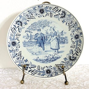 May include: A vintage ceramic plate with a blue and white Dutch scene. The plate features a floral border and a central image of two people, geese, and a windmill. The plate is displayed on a brass stand.