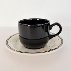 May include: A black ceramic coffee cup with a matching saucer. The cup has a glossy finish and a rounded handle. The saucer is white with black speckles and a black border. The set is designed for serving coffee or tea.