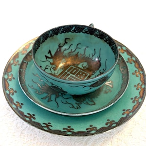May include: A teal teacup and saucer set with a dragon motif. The cup and saucer have a gold and black design. The set is on a white lace tablecloth. The teacup is approximately 2 inches tall and 4 inches wide.