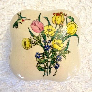 May include: A ceramic trinket box with a floral design. The box is off-white and features a bouquet of yellow and pink flowers, blue accents, and a hummingbird. The box has a unique, rounded shape.