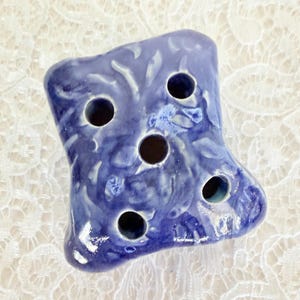 Puede incluir: Un ranero de flores de cerámica azul hecho a mano con seis agujeros. La pieza decorativa tiene una superficie texturizada y un acabado brillante. La herramienta para arreglos florales está diseñada para sujetar los tallos en su lugar.