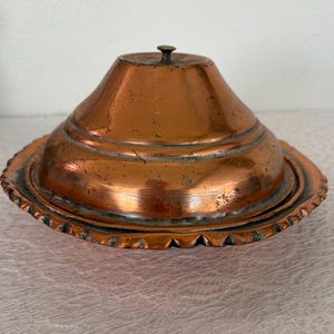 May include: An antique copper butter dish with a domed lid and a decorative, scalloped edge. The lid has a small knob on top. The dish is made of copper and shows signs of age and use.