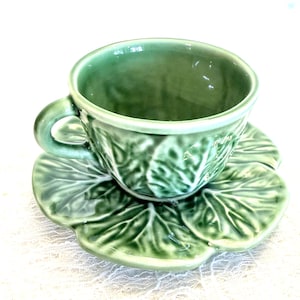May include: A green ceramic teacup and saucer set. The cup and saucer are molded with a leaf design. The cup has a handle and sits on a saucer shaped like a large leaf. The set is a light green color.