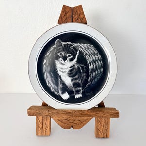 May include: A decorative plate featuring a black and white illustration of a kitten. The plate has a white rim with a black border and is displayed on a small, carved wooden stand. The kitten is the focal point, with detailed shading.