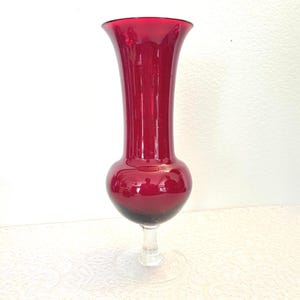May include: A ruby red glass vase with a bulbous base and a flared top. The vase sits on a clear, circular base, and the stem is also clear. The vase is approximately 25 cm tall.
