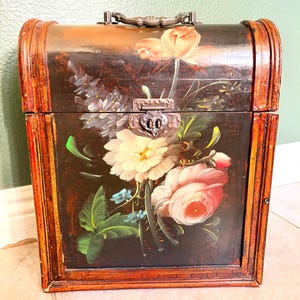 May include: A wooden chest with a dark brown background and a floral painting on the front. The flowers are in shades of pink, white, and blue. The chest has a metal handle and a latch.