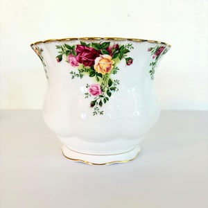 May include: A white porcelain planter featuring a scalloped rim with gold trim. The exterior is decorated with a floral pattern of red, pink, and yellow roses with green leaves. The vessel has a rounded, bulbous body and a flared, wavy top edge.