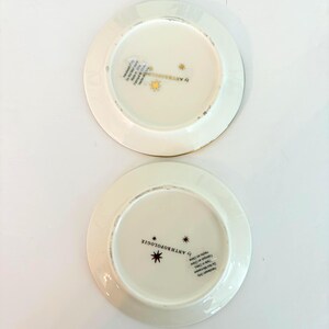 May include: Two off-white ceramic plates with gold rims. The plates have a starburst design and the text "by ANTHROPOLOGIE" printed on the bottom. The plates are on a white surface.