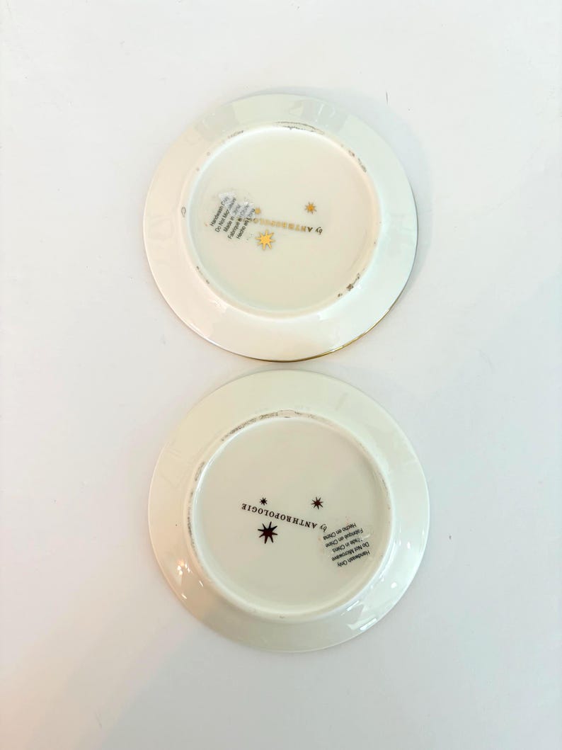 May include: Two off-white ceramic plates with gold rims. The plates have a starburst design and the text "by ANTHROPOLOGIE" printed on the bottom. The plates are on a white surface.
