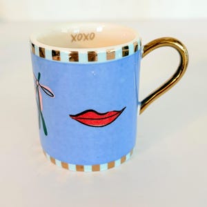May include: A small, light blue ceramic mug with a gold handle and rim accents. The mug features a red lip graphic and the word "XOXO" inside the rim. A pink bow and green scissors are also on the mug.