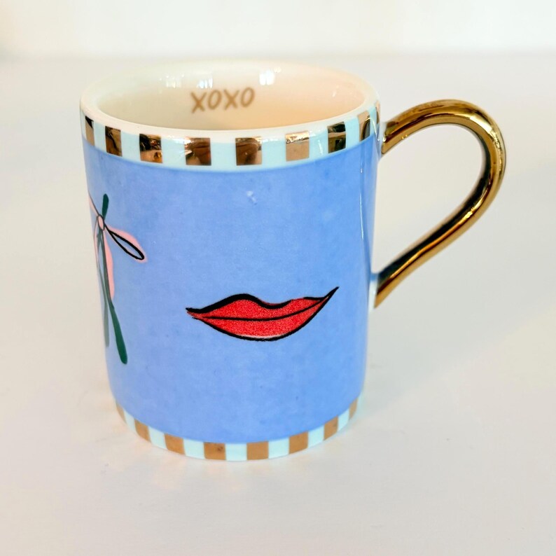 May include: A small, light blue ceramic mug with a gold handle and rim accents. The mug features a red lip graphic and the word "XOXO" inside the rim. A pink bow and green scissors are also on the mug.