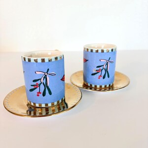 May include: Two ceramic espresso cups with matching saucers. The cups are light blue with a mistletoe design and gold trim. The saucers are gold. The cups and saucers are placed on a white surface.