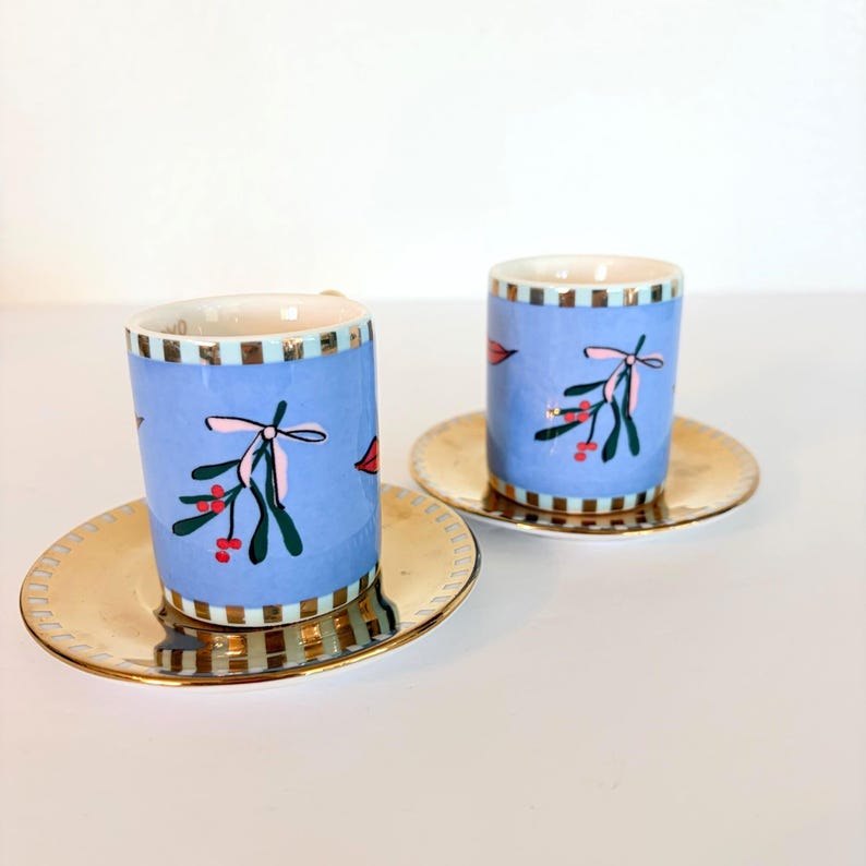May include: Two ceramic espresso cups with matching saucers. The cups are light blue with a mistletoe design and gold trim. The saucers are gold. The cups and saucers are placed on a white surface.