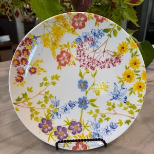 May include: White ceramic plate with a colorful floral design. The plate features a variety of flowers in shades of red, yellow, blue, and purple, with green leaves and stems. The plate is displayed on a black metal stand.