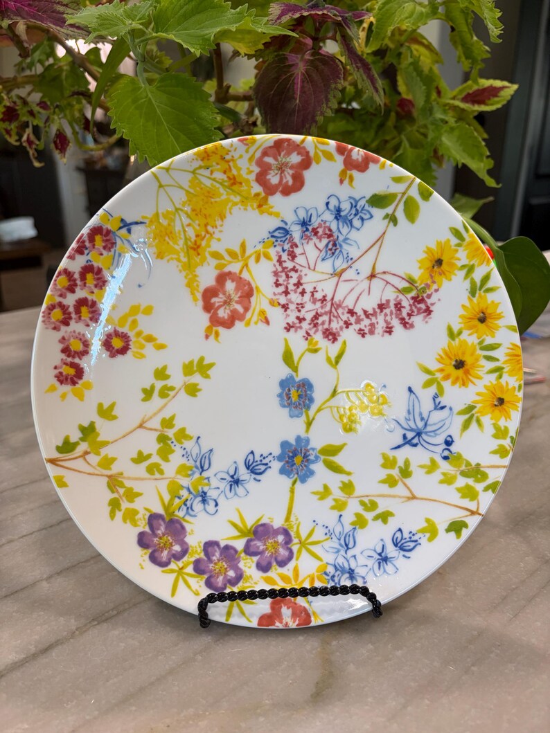 May include: White ceramic plate with a colorful floral design. The plate features a variety of flowers in shades of red, yellow, blue, and purple, with green leaves and stems. The plate is displayed on a black metal stand.