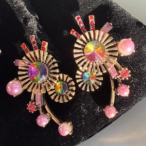 Könnte beinhalten: Ein Paar goldfarbene Statement-Ohrringe mit Sonnenstrahlen-Design. Jeder Ohrring hat einen großen, schillernden Mittelstein, umgeben von strahlenden goldenen Strahlen, rosa und roten Akzenten und rosa perlenartigen Perlen. Die Ohrringe sind vor einem schwarzen Hintergrund platziert.