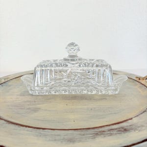 Op de afbeelding: Een heldere, rechthoekige glazen botervloot met een deksel. De schaal heeft een decoratief, gestructureerd patroon en een kleine handgreep aan de bovenkant. De schaal staat op een ronde, lichtgekleurde, verweerde schaal.