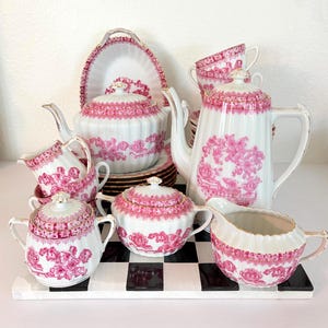 May include: A white porcelain tea set featuring a pink transferware pattern of floral and landscape motifs with gold trim. The set includes a tall teapot, a lidded sugar bowl, a creamer, teacups, saucers, and a serving platter with fluted edges.