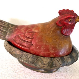 May include: A hand-carved wooden chicken figurine, painted in shades of red, brown, and green. The rooster sits atop a decorative, weathered-looking nest. The figurine is a decorative item, likely for a kitchen or farmhouse-style decor.
