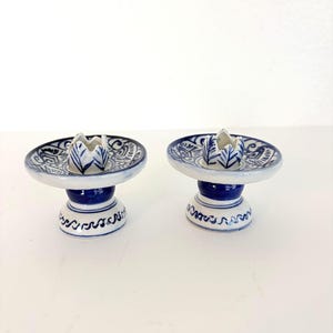 May include: Two matching ceramic candle holders. Each has a white base with blue accents and a blue and white patterned top. The candle holders have a small, white, flower-shaped design in the center.