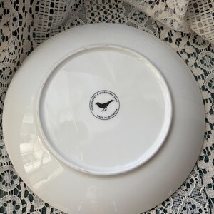 May include: White ceramic plate with a circular black graphic of a bird and the text "Dishwasher & Microwave Safe" and "Made in Indonesia". The plate is on a white lace surface.