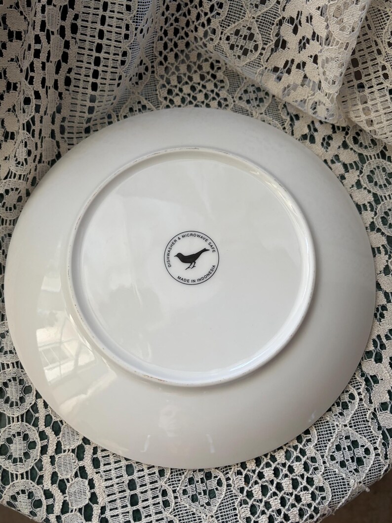 May include: White ceramic plate with a circular black graphic of a bird and the text "Dishwasher & Microwave Safe" and "Made in Indonesia". The plate is on a white lace surface.