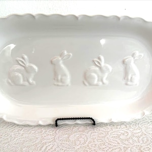 May include: A white ceramic mug with a black handle and the text "But First, Coffee" in a bold, black font. The mug is set against a blurred background, suggesting a cozy setting.