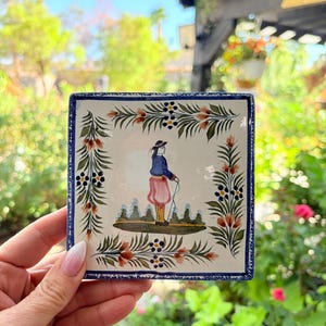 Könnte beinhalten: Eine quadratische Keramikfliese mit einer handgemalten Illustration einer Figur in blauer Jacke und rosa Hose, die in einer Landschaft steht. Das zentrale Bild ist von einem dekorativen Rand aus grünen Blättern, orangefarbenen Blumen und blauen Beeren umrahmt.