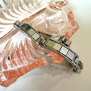 May include: A silver-toned bracelet with square, mirrored segments rests on a pink glass dish. The dish has a floral design. The bracelet's clasp is visible, and the overall aesthetic is vintage.