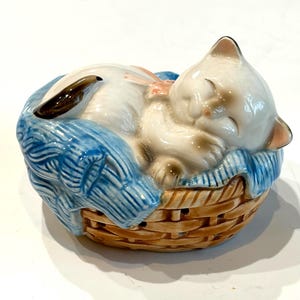 Puede incluir: Figura de cerámica de un gato durmiendo en una cesta tejida. El gato es blanco con detalles marrones, descansando sobre una manta azul tejida. La cesta es marrón con una textura tejida.