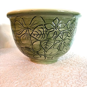 Può includere: Una ciotola in ceramica verde con un motivo floreale. La ciotola ha una forma ampia e arrotondata ed è decorata con fiori e foglie in rilievo. L'interno della ciotola è di una tonalità di verde più chiara. La ciotola è su una superficie di pizzo bianco.