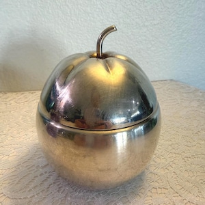 May include: A silver-colored apple-shaped container with a lid and a curved stem. The container has a smooth, reflective surface and sits on a white lace tablecloth. The lid fits snugly on the base, creating a seamless look.